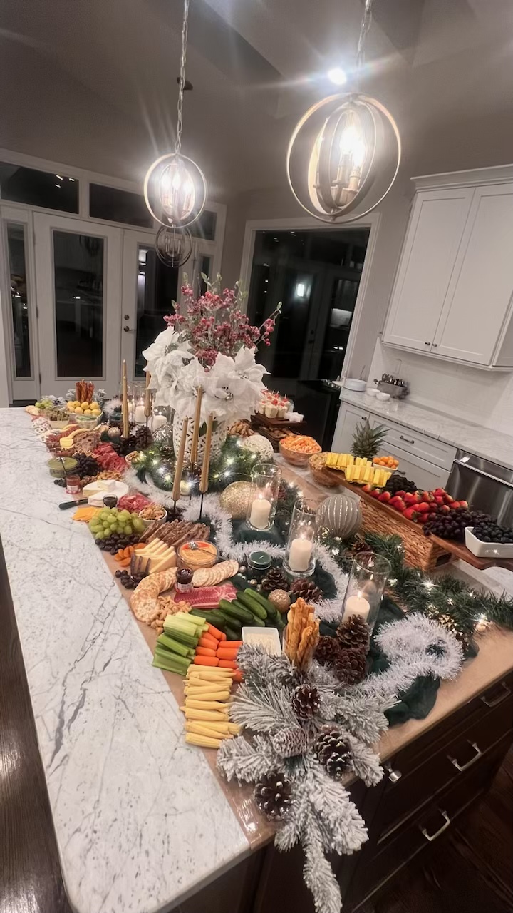 A grand designed grazing table on a marble island, styled with white poinsettias, flocked greenery, candles, and a curated charcuterie spread.